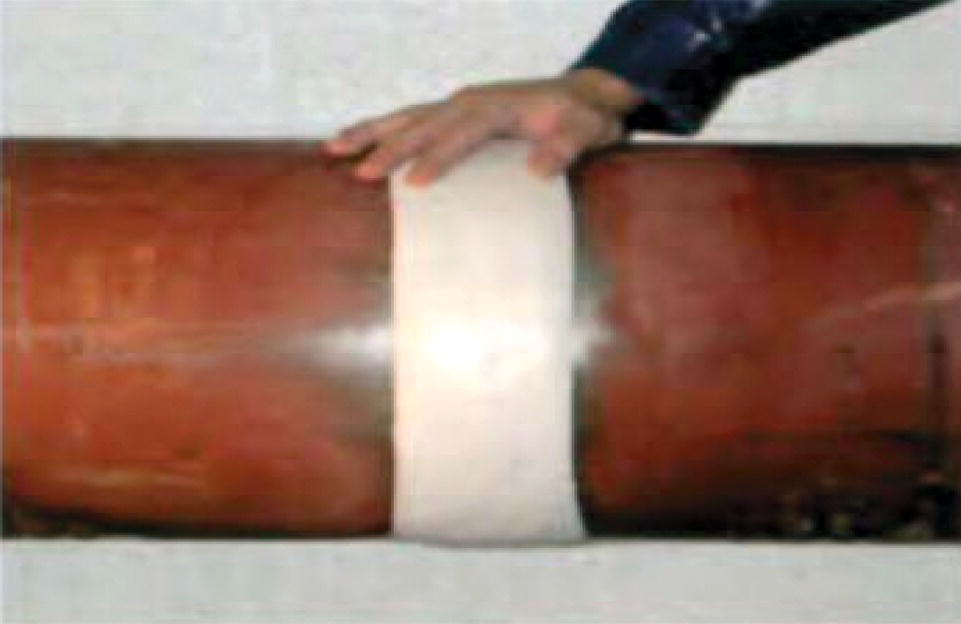 A photograph of a pipe with a repair tape or wrap being applied. A person’s hand is visible at the top of the frame, pressing down on a light-coloured repair tape or bandage that encircles the dark pipe. The tape creates a distinct band around the pipe’s circumference, positioned at what might be a joint or damaged area. The pipe runs horizontally across the frame, and the contrast between the dark pipe surface and lighter repair material is clearly visible.