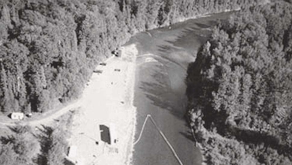 An aerial photograph captures a curved river or waterway flowing through a forested landscape. The water forms a distinct contrast with the surrounding terrain. On one side of the waterway is a light-coloured sandy or gravelly beach area with a small structure or object. A thin line, possibly a rope or cable, extends from the shore into the water. Dense trees and vegetation border both sides of the waterway, creating a natural corridor through the wilderness. The perspective provides a clear view of how the waterway bends through the forest environment.