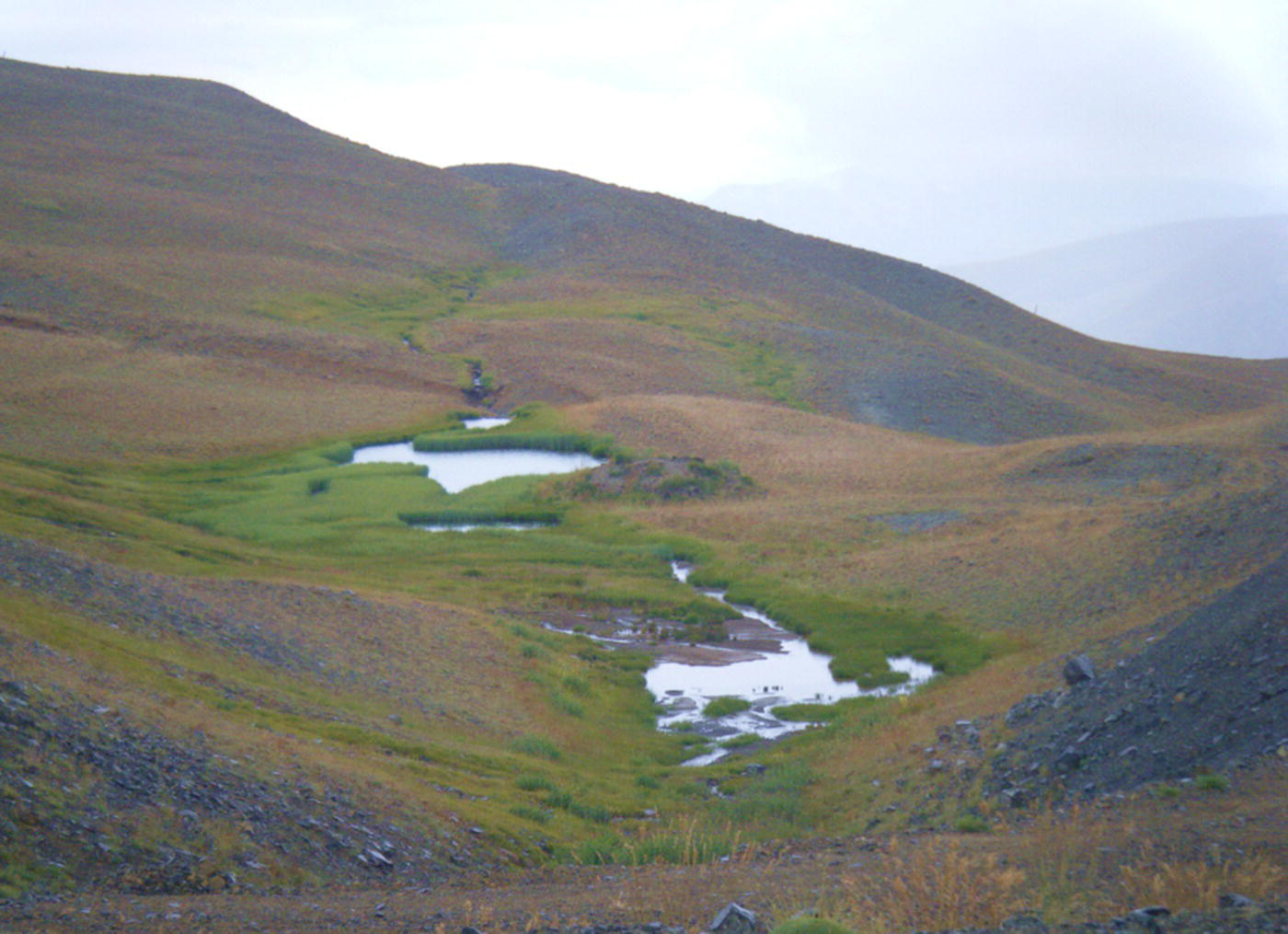 A photograph captures a barren mountainous landscape with gentle rolling hills. A narrow valley runs through the centre, containing small bodies of water that appear as patches against the darker terrain. A stream or watercourse connects these water bodies, flowing down the valley floor. The hillsides have minimal vegetation and display erosion patterns. The topography creates a natural basin or watershed area. The sky is light-coloured and hazy, creating contrast with the darker hills.