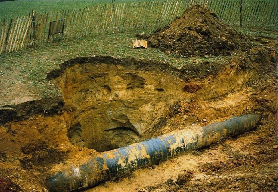 A photograph presents an excavation site with an exposed underground pipeline. A large cylindrical pipe runs horizontally across the bottom portion of the image. Above the pipe is a significant hole or trench with clearly visible soil layers and stratification in the excavated wall. A pile of excavated dirt sits on the right side of the trench. The surrounding ground appears disturbed and muddy. In the background, there are some fencing or barriers. The image captures a utility work, pipeline maintenance, or archaeological excavation revealing subsurface infrastructure.