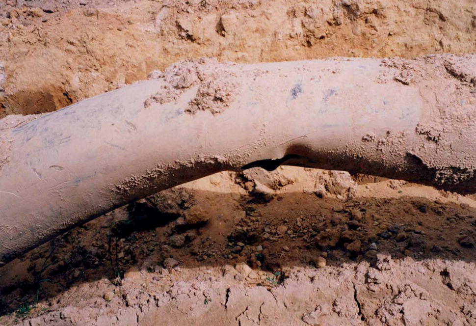 A close-up of a pipeline damage reveals intricate textures and surface details, highlighted by the contrasts in monochrome. Surrounding elements include sand blanketing the ground. Shadows cast by the rock enhance its prominence against the soft backdrop, emphasizing the rugged features and natural beauty.