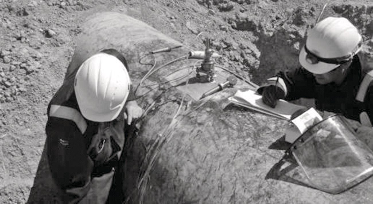 A group of men wearing helmets examines a map, indicating an emphasis on teamwork and planning. Dressed in workwear suitable for outdoor environments, they appear to be engaged in a construction or engineering project. The image features a monochromatic palette, emphasizing the seriousness of their task against a natural backdrop. The presence of hard hats highlights the collar nature of their profession while reinforcing safety measures associated with construction work.
