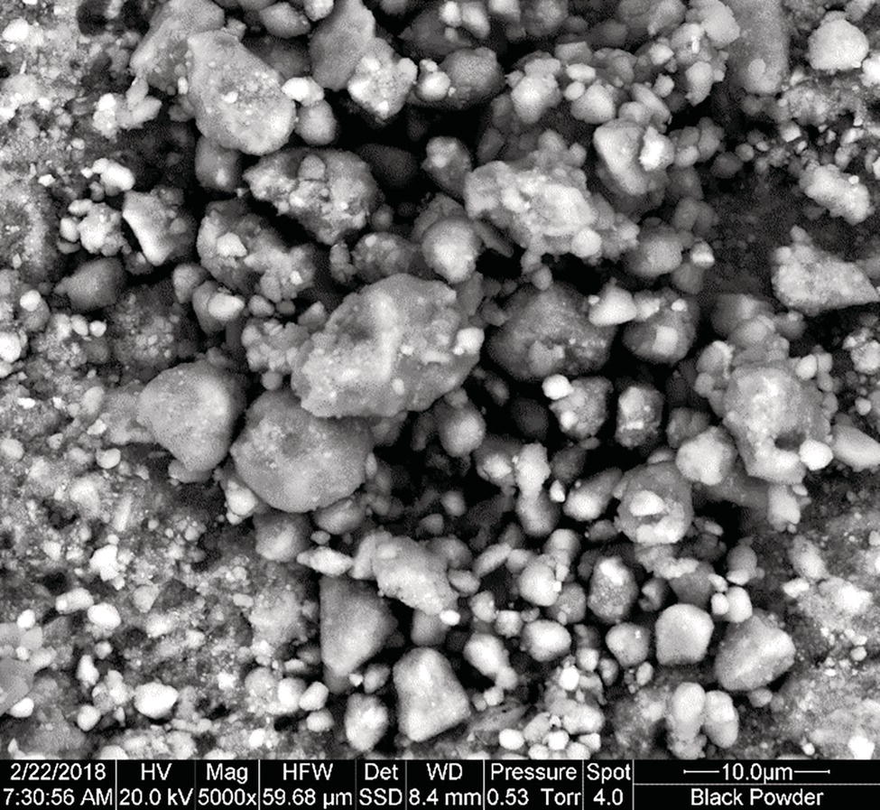 A microscopic photograph showing clusters of black powder particles with a rough texture. The image has a scale bar indicating 10 micrometers, captured at 5000x magnification.