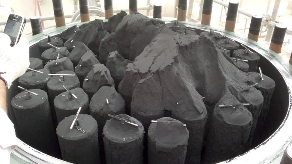 A top-down view of a circular setup filled with dark powder, revealing several vertical rods protruding from the surface. A hand holding a phone is visible on the left side of the image, indicating scale and context.
