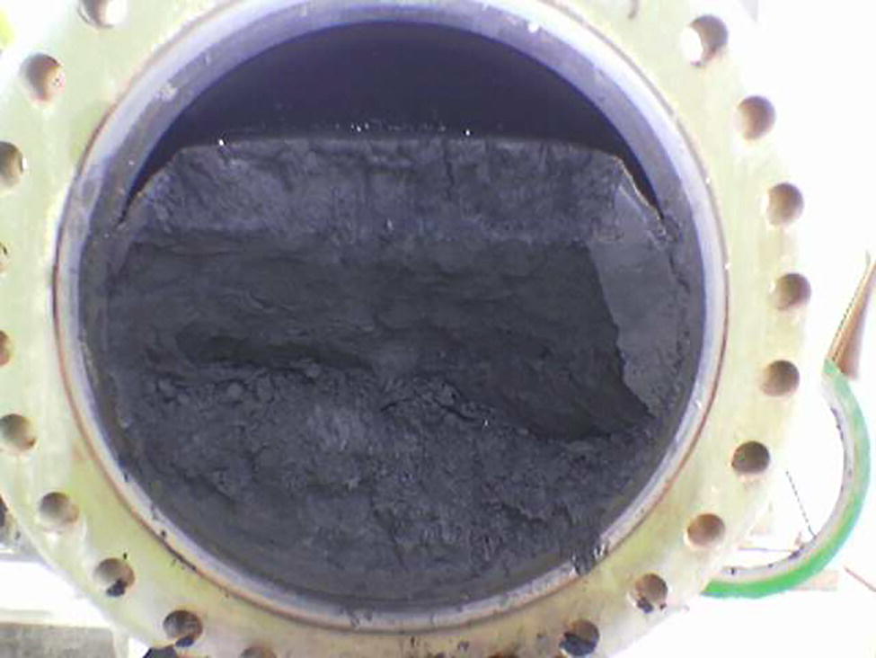 An interior view of a cylindrical vessel showing accumulated dark sludge or sediment at the bottom. The image highlights the vessel’s circular rim with visible bolt holes and a rough surface texture.