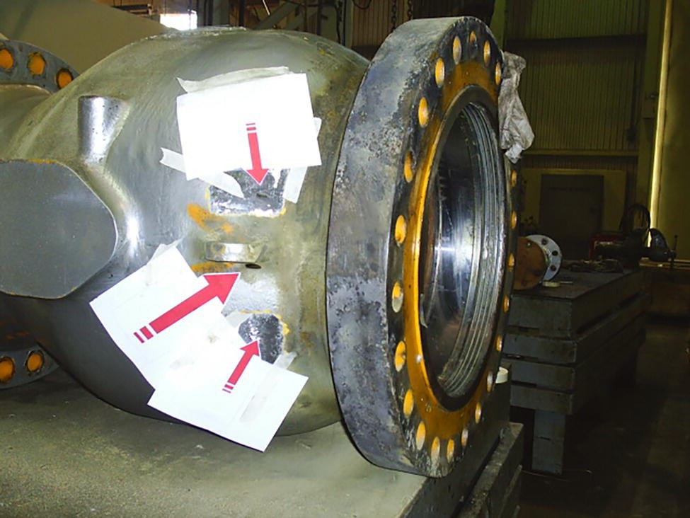 A photograph of a large industrial valve with white paper labels indicating points of interest. The valve is positioned on a work surface in a workshop setting.