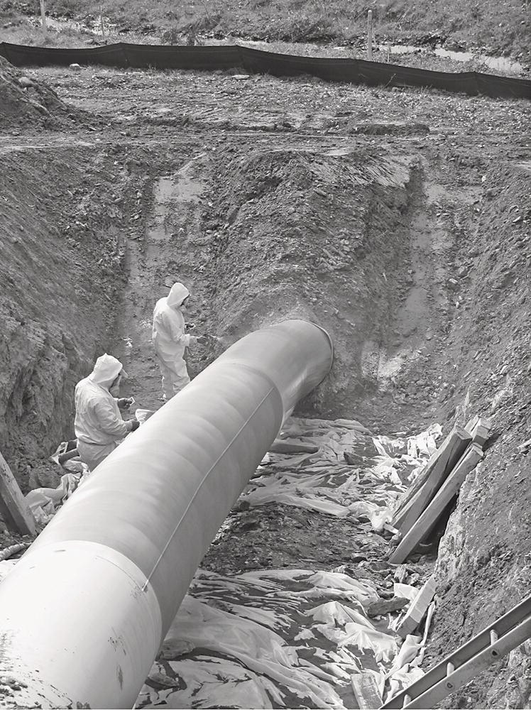 A photograph shows workers in protective suits inspect a large pipeline laid in a trench. The surrounding area shows freshly dug earth and plastic coverings for protection.
