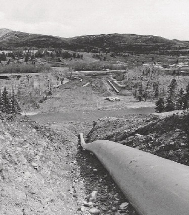 An image of a large pipeline running through a hilly landscape. The background features a valley with several additional pipes visible and mountains in the distance.