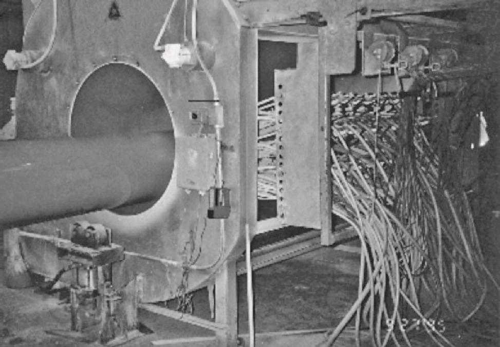 A photograph showing a large pipeline entering a testing chamber. Numerous cables and connections are visible on the side, indicating equipment for monitoring or processing.