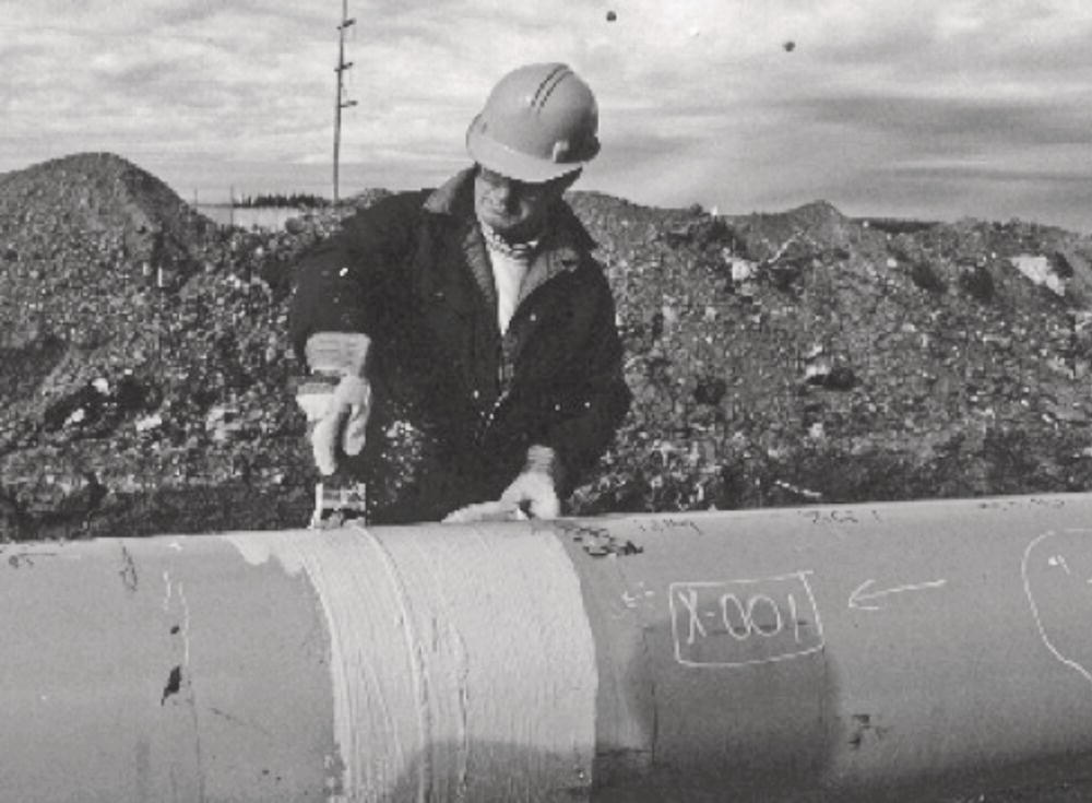 A photograph of a worker in a hard hat applies a coat of material to a large pipe using a brush. The background shows a construction site with debris and a cloudy sky overhead.