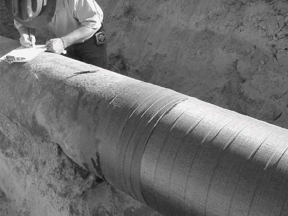 A worker examines a large pipe covered with protective material while taking notes. The pipe is partially buried in the ground, with sand surrounding the area.