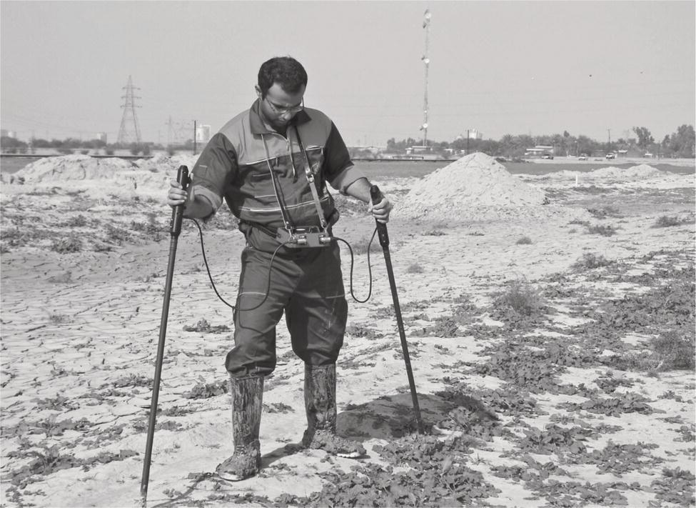 A surveyor wearing boots and a harness uses two probes to assess ground conditions. He stands on a sandy area with piles of material in the background.