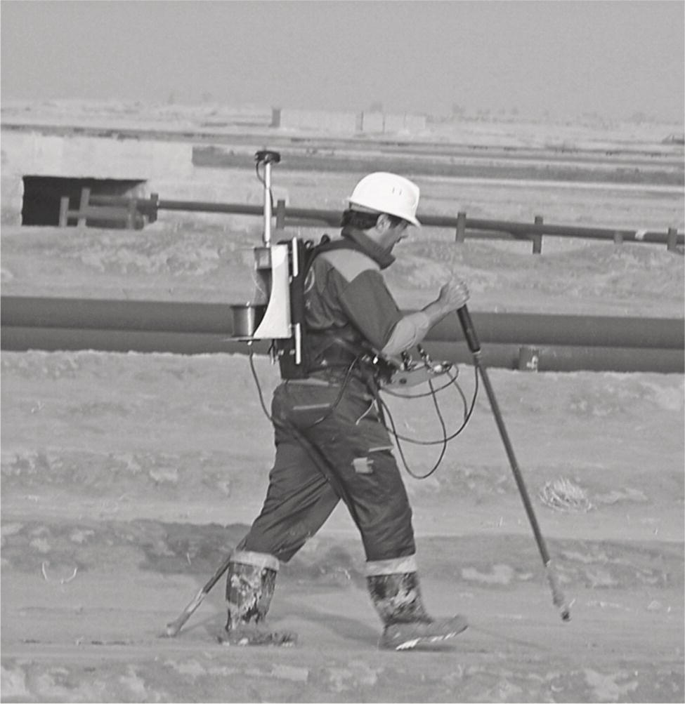 A surveyor wearing a safety helmet and harness walks across a sandy area, carrying equipment on his back. He uses a probing stick to assess the ground near pipelines in the background.