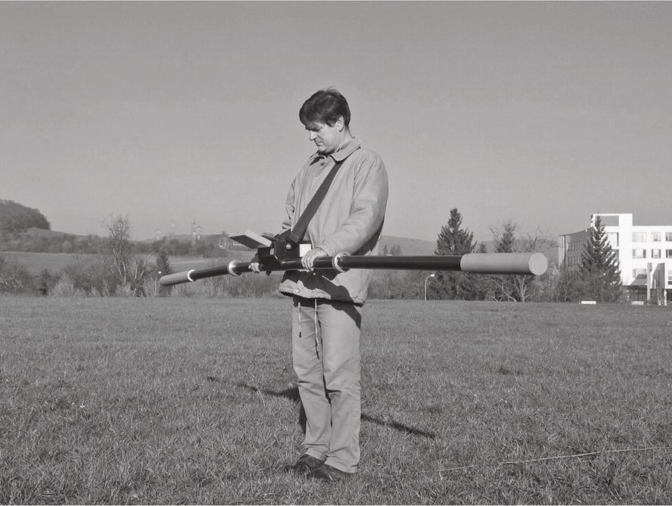 A photograph of a person stands on a grassy field, holding a surveying instrument with long rods. In the background, a building and trees are visible under a clear sky.