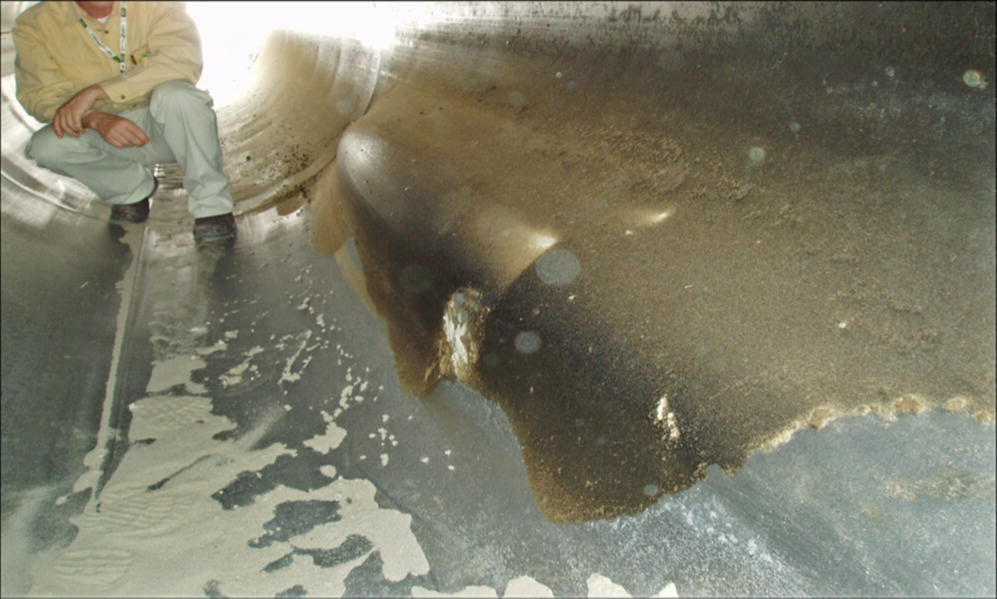 An interior view of a pipeline with a worker sitting nearby. Water flows along the surface, highlighting a damaged section of the pipe.