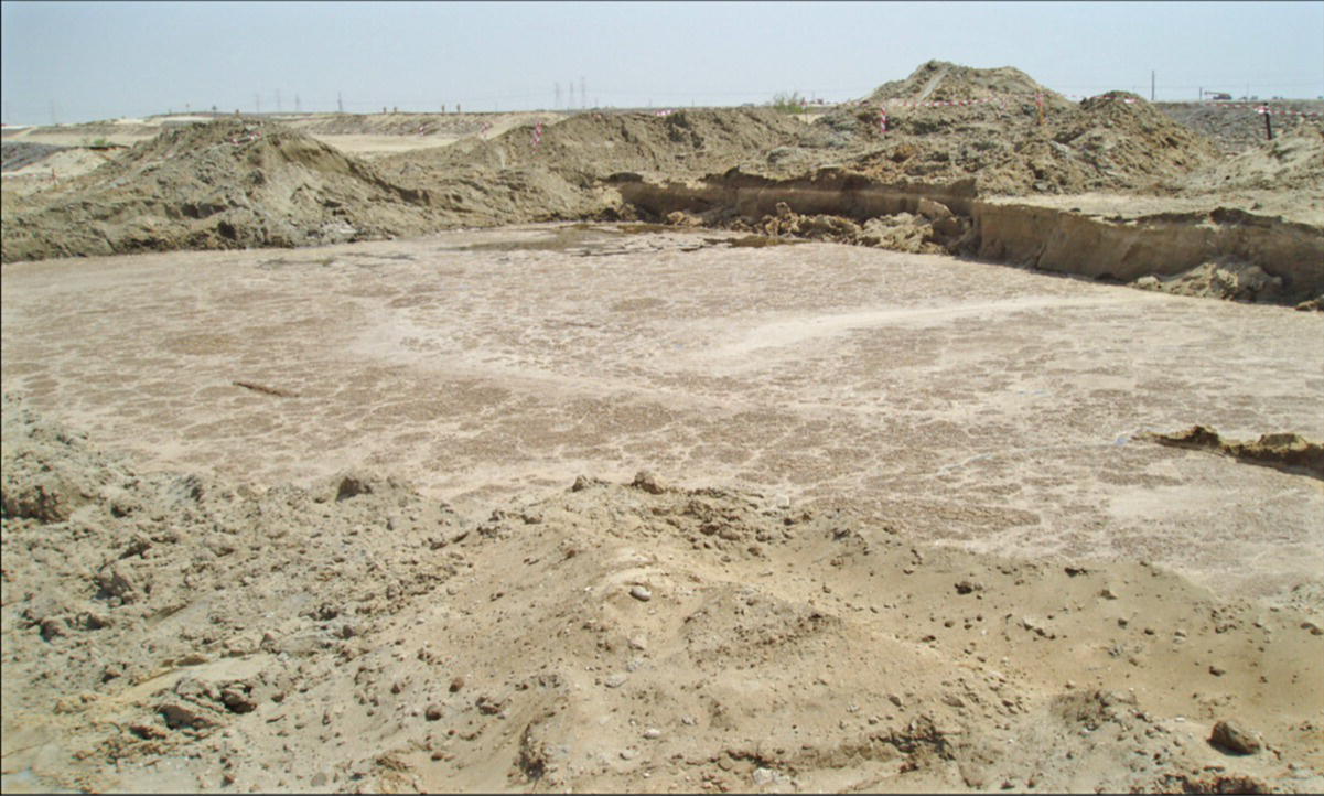 A large, muddy area with water pooling in the center, surrounded by rough terrain. The scene appears barren with hills of sand and gravel in the background.