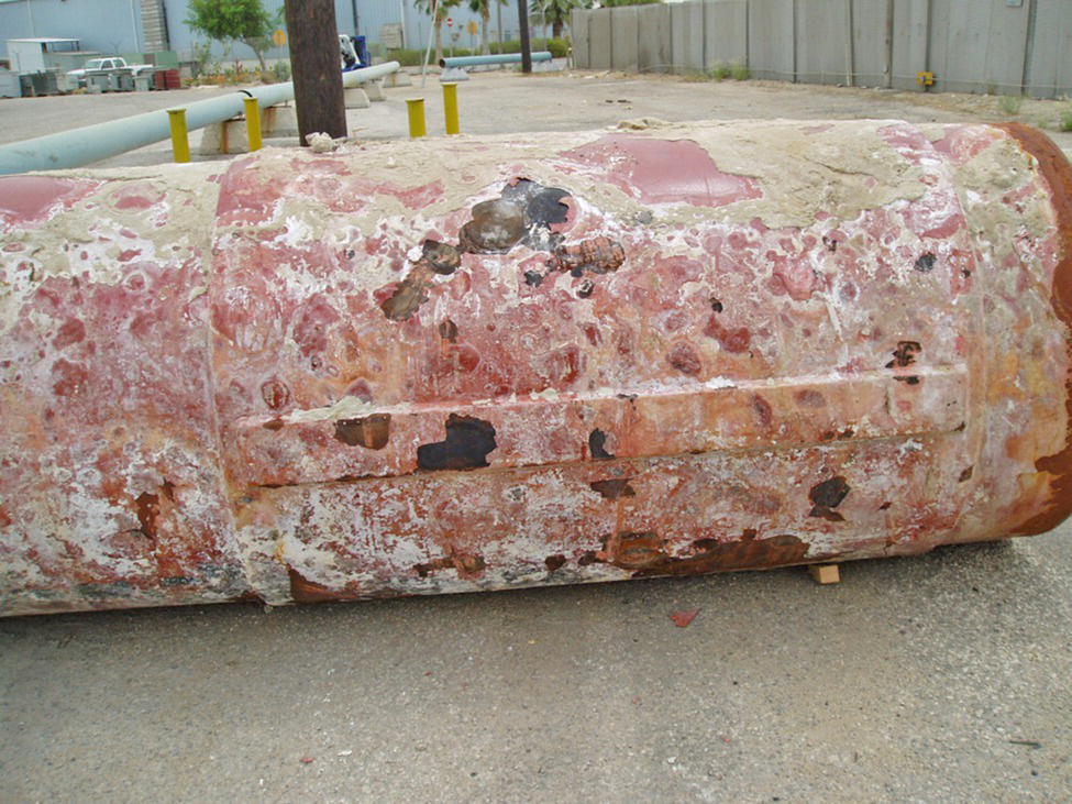 A large, weathered cylindrical object resting on the ground, featuring significant surface corrosion and discoloration. The background shows a barren area with a faint outline of structures.