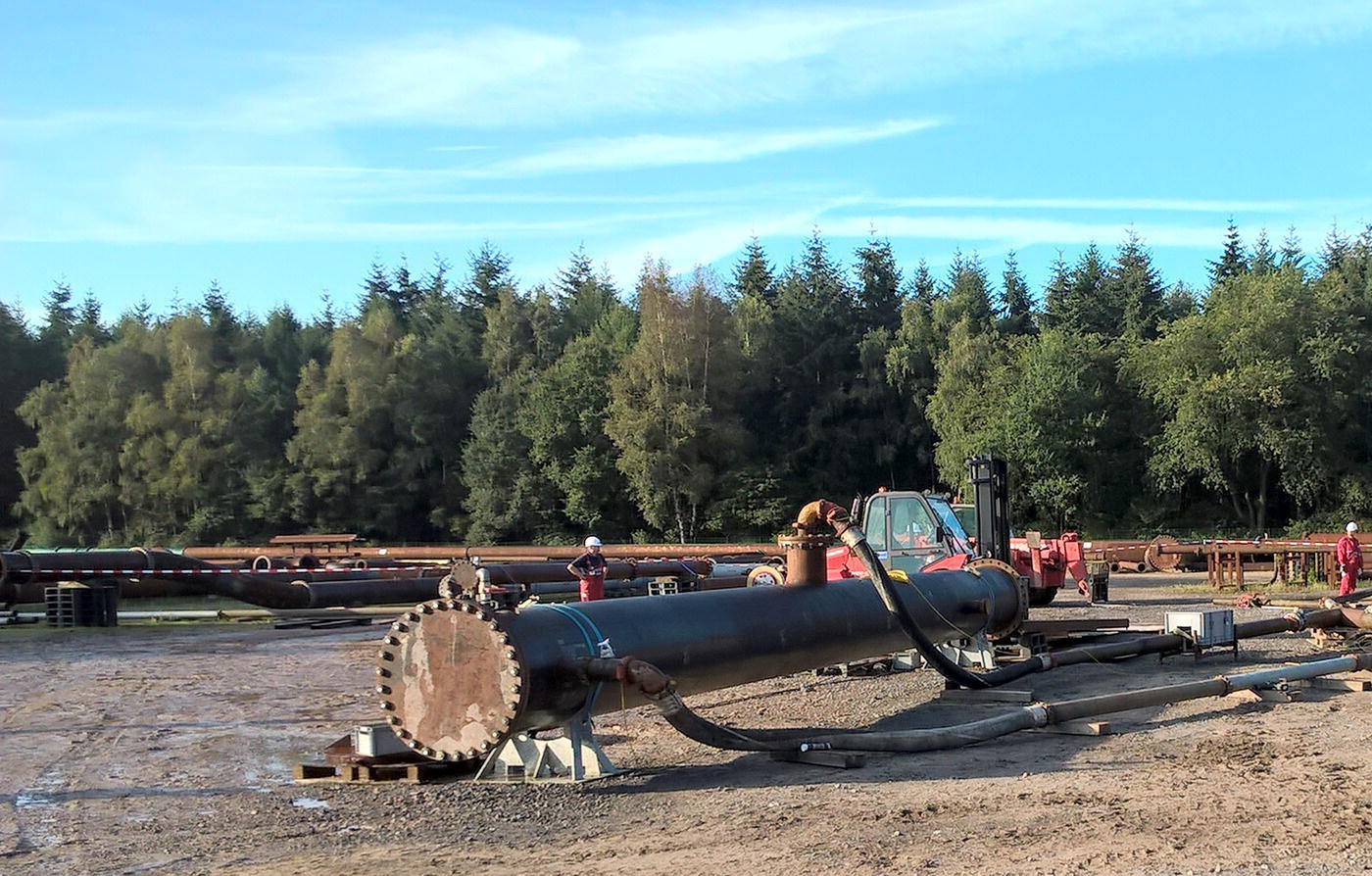 An industrial site with large pipelines and machinery set against a backdrop of trees. The ground is bare, indicating ongoing construction or installation activities.